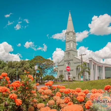 Paróquia São José em Boa Vista do Buricá, Rio Grande do Sul, em um dia ensolarado com céu azul turquesa e poucas nuvens brancas. Em primeiro plano, um jardim bem cuidado com grama aparada e flores laranja vibrantes.