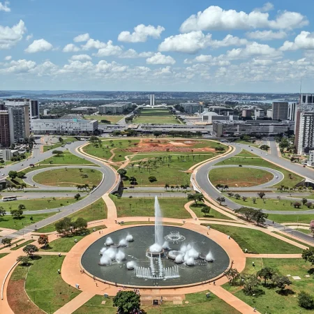 Vista aérea da Esplanada dos Ministérios em Brasília durante o dia, com a Torre de TV ao fundo e a Fonte da Torre de TV em primeiro plano. A área é bem arborizada, com um extenso jardim bem aparado, árvores e ipês cor-de-rosa floridos. Poucos veículos são visíveis entre os prédios modernos e a arquitetura limpa.